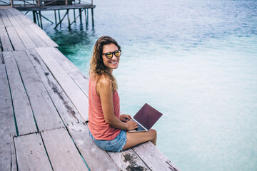 Smiling female sitting on pier and typing on notebook