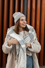 Joyful young woman model in a stylish knitted hat in a glamorous faux fur coat with a woolen warm...