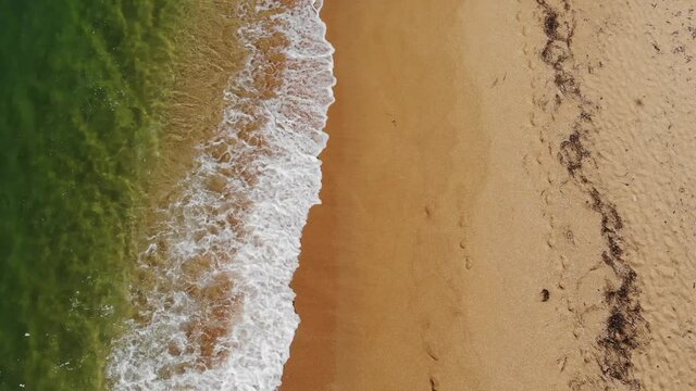 Aerial view close-up low flight over sea waves rolling on the golden sandy coast. Calm surf behind the seashell beach
