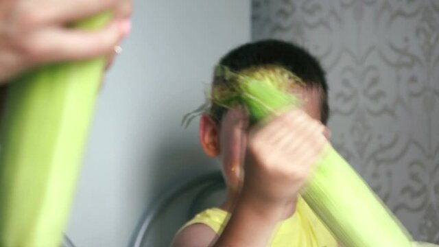 Cheerful Caucasian boy sitting at the table in the kitchen peels ripe sweet yellow corn. Fresh harvest of corn. Selective focus, shallow depth of field. Child's face out of focus