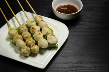 Grilled pork meatballs skewer in a white plate and sauce in a white bowl on a black background.