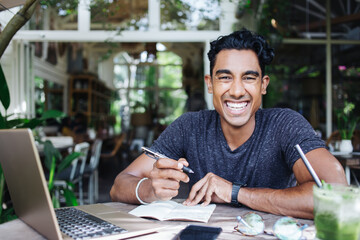 Cheerful adult ethnic man looking at camera while sitting and writing in notepad in cafe