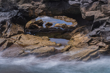 A stone arch by the sea displays calm blurred water and rocks behind.