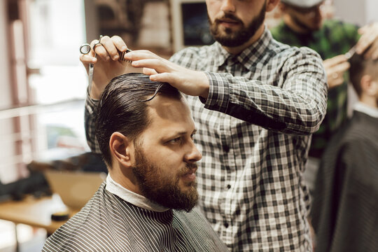 Haircut in the barbershop. Close up view of young bearded man getting haircut while sitting in chair at barbershop. Hairdresser cuts hair with scissors and a comb