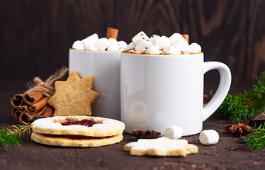 Two cups of hot chocolate or cocoa with marshmallow and cinnamon stick, bitter chocolate, winter spices and jam filling cookies. Festive Christmas or New Year decoration. Dark stone background.