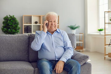 Fototapeta premium Senior man sitting on couch and making phone call on smartphone to keep in touch with his relatives