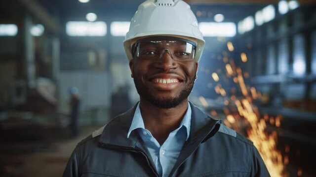 Happy Professional Heavy Industry Engineer/Worker Wearing Uniform, Glasses and Hard Hat in a Steel Factory. Smiling African American Industrial Specialist Standing in a Metal Construction Manufacture.