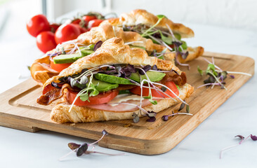 Three croissant sandwiches on a wooden board against a sunny window with small vine tomatoes in soft focus in behind.