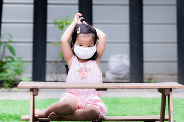 Fototapeta na wymiar Child wearing white cloth mask sits on wooden table. Children wear pink clothes. Kid use both hands to pull the elastic strap of the cloth mask. Prevent the spread of viruses and dust pm 2.5.