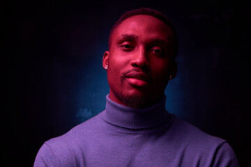 portrait photo on black background with neon light on dark-skinned handsome man in a blue turtleneck who is looking at you