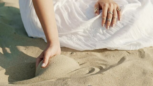 Woman hands playing with sand at beach. Beautiful scene of young boho gypsy girl in white dress with rings touching grit. Slow motion