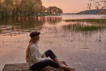 A barefoot girl with long blonde hair and a cap sits on the bridge by the lake against the...