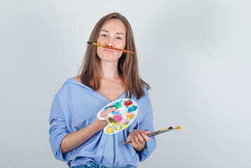 Young woman in shirt, shorts holding paint brush and palette and looking funny , front view.