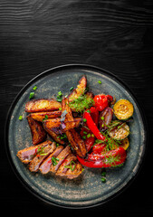 fried meat with potatoes, pepper, tomatoes, herbs and spices in plate on black wooden table background