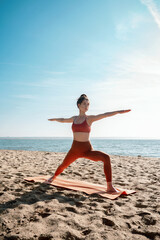 Fototapeta na wymiar Young adult female practising yoga on a beach, Virabhadrasana II pose