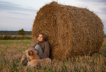 A teenage girl hugs a dog in a field while sitting by a roll of hay. Autumn calm landscape.