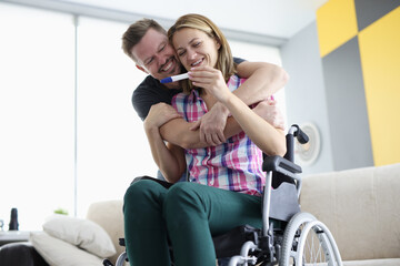 Husband hugs his wife in wheelchair by shoulders with positive pregnancy test. Disabled women...