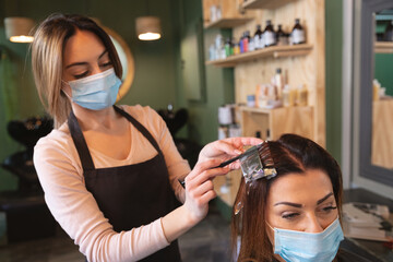 Female hairdresser wearing face mask dying hair of female customer at hair salon