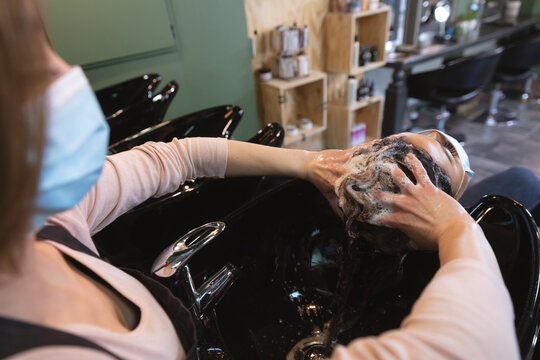 Female hairdresser wearing face mask washing hair of female customer at hair salon