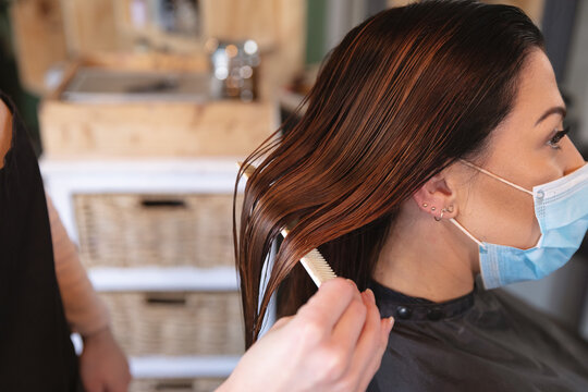 Female hairdresser combing hair of female customer wearing face mask at hair salon