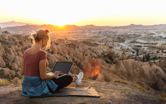 Young girl working at laptop at sunset on top of mountain with beautiful view at valley