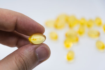 One yellow capsule that is held in fingers on a white background. The background is blurred on which is a group of yellow capsules. Vitamins