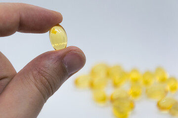 In the foreground is a yellow capsule that is held in the fingers. In the background is a group of yellow capsules. The background is blurry. White background