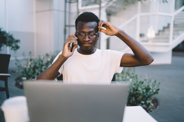 Serious african american male entrepreneur checking banking account on laptop computer making mobile phone call, pensive dark skinned man freelancer confirming online booking via cellular talk