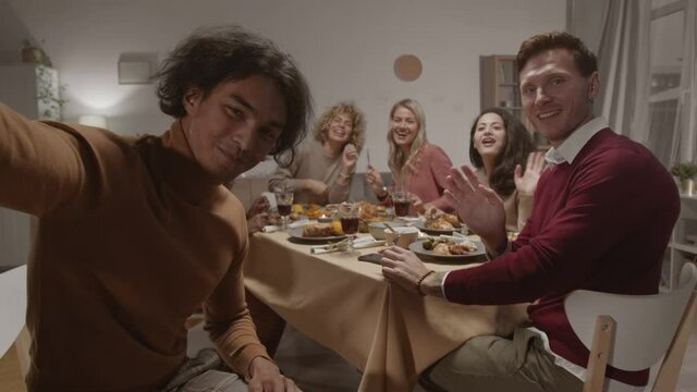 Medium shot of young mixed-race man holding camera in his hands and making selfie with his multiethnic friends sitting at table and having festive dinner