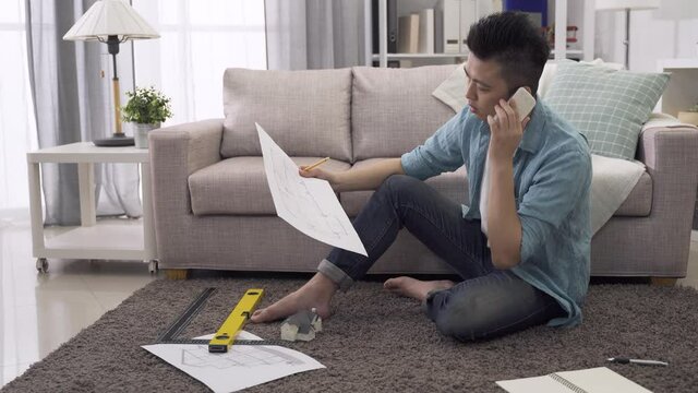 seated on rug surrounded by measuring tool and prints, male architect looking at construction plan and house miniature while having phone discussion at home.