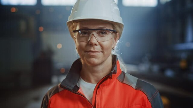 Portrait of a Professional Heavy Industry Engineer/Worker Wearing Uniform, Glasses and Hard Hat in a Steel Factory. Beautiful Female Industrial Specialist Standing in Metal Construction Facility.