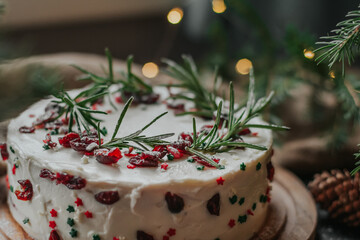 Christmas cake with white cheese cream, decorated with cranberries and rosemary. New Year's treat on the background of fir branches and garlands.