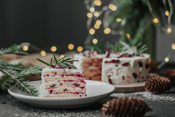 A piece of Christmas cake on a white plate against the background of fir branches and garlands. Cranberry pie with white cream, decorated with rosemary.