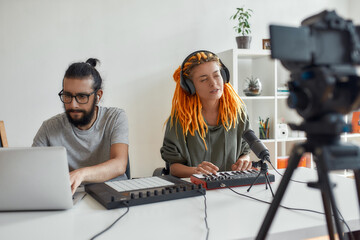 Woman with dreadlocks singing and playing. Female and male blogger making music using synthesizer, drum pad machine and laptop, recording video blog or vlog at home