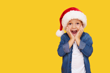 Portrait of a satisfied little child boy dressed in christmas Santa hat. laughing isolated over colored yellow background. Wow face. Season of discounts