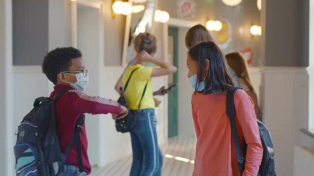 Side view of diverse schoolkids in facial mask greeting with elbow bump in school corridor