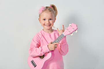 Cute caucasian little girl 4-5 year old in pink sweatshirt are playing the ukulele against gray studio background
