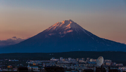 View of the volcano at sunset. The top of the volcano is illuminated by the sun. In the foreground is the evening city