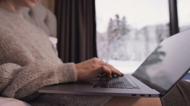 close-up of the girl hands print something on the laptop sits on a bed with a laptop in her hands against the panoramic windows behind which the winter forest in the snow.