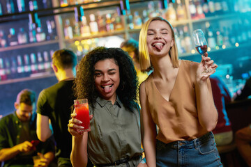 Attractive young women getting drunk, showing tongue to camera, posing with cocktail in their hands. Friends celebrating, having fun in the bar