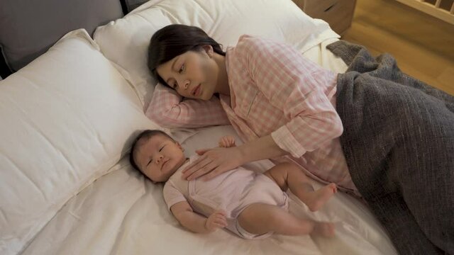 asian mother lying down with her lovely newborn on bed at night is getting her child to sleep by touching and talking to her softly at home.