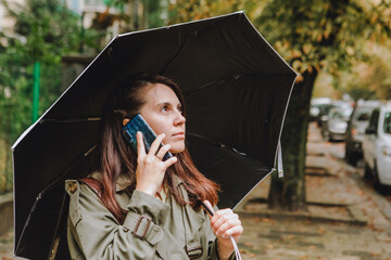 young pretty woman talking on the phone at city street hiding from rain under umbrella