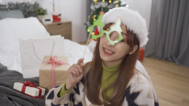 attractive asian woman with Christmas hat looking at camera is making a face by pursing lips and crossing her eye while holding stick mask and artificial mustache.