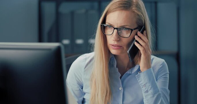 Attractive business lady in eyeglasses and blue shirt looking at computer screen during mobile conversation. Confident woman with blond hair sitting at office and using modern technology.