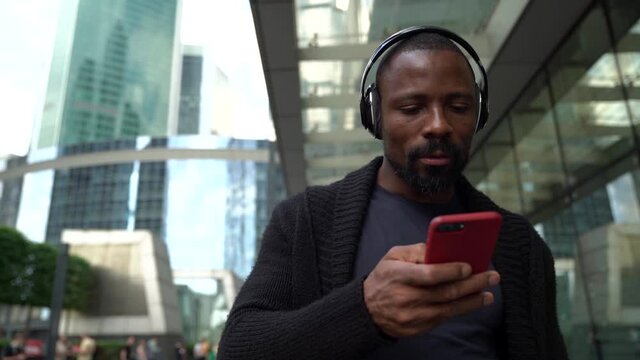 An inspired cheerful black man sings and listens to music in wireless headphones and a smartphone on a city street in the background of business centers.