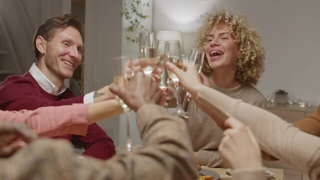 Back view of several cheerful people sitting at dining table with champagne glasses in their hands, clinking them, making a sip while chatting
