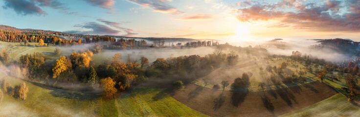 Stunning aerial panorama of a misty rural landscape at sunrise, with colorful autumn trees, fields and meadows, blue sky, trees casting shadows and beautiful wafts of mist