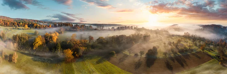 Atemberaubendes Luftpanorama einer nebligen ländlichen Landschaft bei Sonnenaufgang, mit bunten Herbstbäumen, Feldern und Wiesen, blauem Himmel, Bäumen, die Schatten werfen, und wunderschönen Nebelschwaden © Smileus