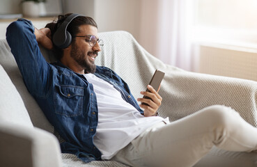 Relaxed indian guy enjoying listening music on smartphone with wireless headphones
