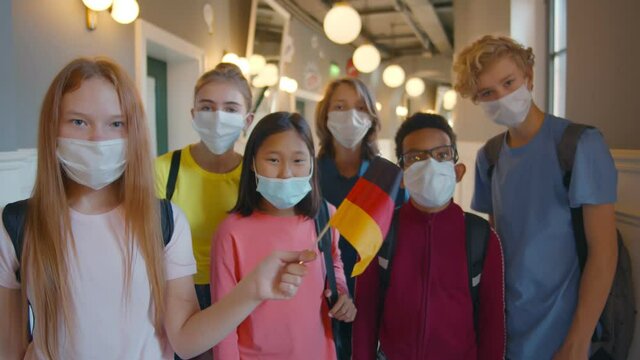 Diverse school students in mask standing in corridor holding german flag
